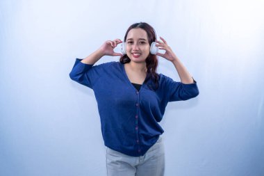 Asyalı Endonezyalı kadın mavi hırka ve kulaklık giyip müzik dinliyor.