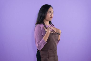 Kahverengi önlük ve mor gömlek giyen genç bir kadın, dramatik bir yüz ifadesi ve göğsünde elleri olan mor bir arka plana karşı duruyor..