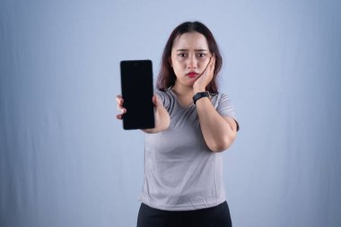 Gri tişörtlü genç bir kadın, yanağını tutarken ve kameraya doğru akıllı bir telefon uzatırken hayal kırıklığına uğramış görünüyor..