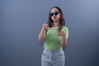 Güneş gözlüğü ve yeşil bluz takan kendine güvenen genç bir kadın, gösterişli hareketler yapar. Düz bir arkaplan önünde cesur bir ifadeyle..