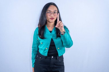 Turkuaz gömlekli ve gözlüklü Endonezyalı bir kadın işaret parmağını kaldırarak uyarı işareti yapıyor..