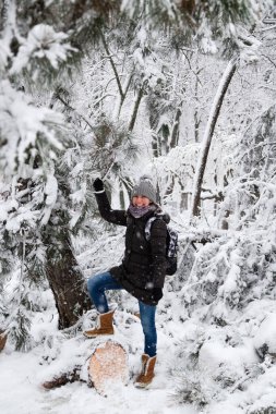 Smiling young woman standing on fallen tree after sleet load and snow in a snow-covered winter park. Girl enjoying snowy winter, frosty day. Walk in winter forest.