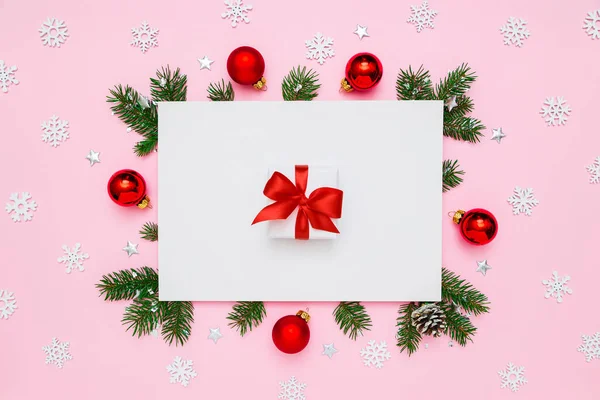 Christmas, New Year holiday layout. White gift box with red bow in frame of natural fir tree branches and decorations on white and pastel pink flat lay background