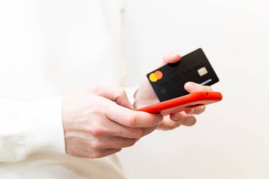 Kyiv, Ukraine - December, 2 2022: Close up of female hands holding Monobank Mastercard credit card and using smartphone in red case on white background. Paying securely online, using banking service.