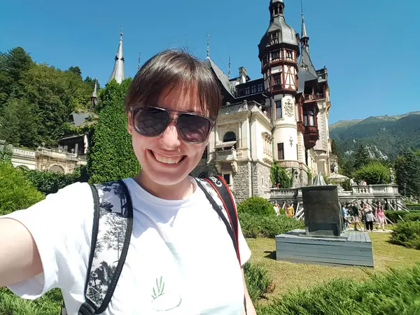 stock image Sinaia, Romania - August 26, 2022: A young, happy, smiling girl with a backpack and camera taking a selfie for social media in front of Peles Castle. Travel Romania concept.