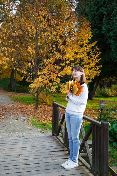 stock image Photo of cute young romantic dark-haired girl in white sweater, jeans, and sneakers, holding bouquet of orange fallen leaves in autumn park. Cute 30s woman, no make up, closed eyes.