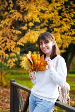 Güz parkında elinde bir buket turuncu yaprak tutan, beyaz kazak ve kot giyen, gülümseyen genç ve güzel kızın portresini yakından çek. Kameraya bakıyorum. Makyajsız.