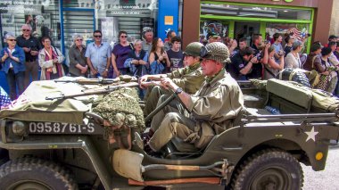 Aziz Michael L 'Eglise, FRANSA - 6 Haziran 2019. Normandiya Çıkarması 'nın kutlanması, silahlı çıkarma, İkinci Dünya Savaşı' nın Normandiya 'da sona ermesi, müttefik ülkelerin yardımıyla. Askerler sokakta izleyicilerle birlikte cipte yürüyorlar.