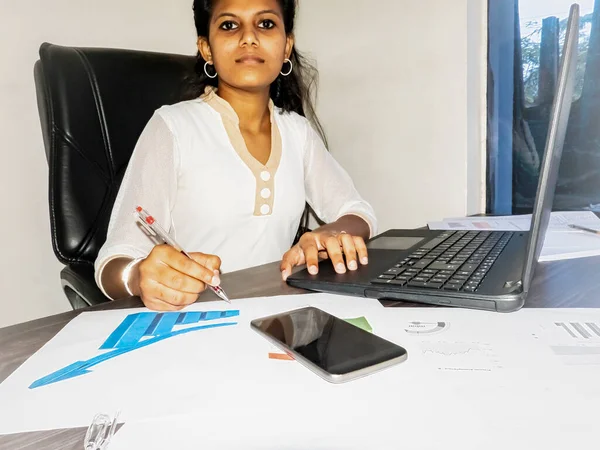 Young indian student adult business woman writting at office indoors with laptop, mobile and sheets with graphs on the desk to illustrate communication and technology concept.