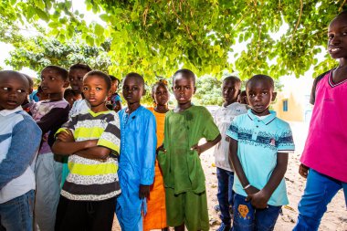 Bour, Senegal - Aralık 2020 civarı. Kimliği belirsiz bir grup genç, güneşli bir yaz gününde okulun bahçesinde ayakta dikiliyorlar. Uluslararası NGO 'nun yeni çantalarını bekliyoruz. Üzgün ve zavallı çocuklar..