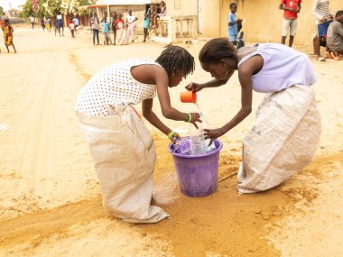 Bour, Senegal, Afrika, Aralık 2021 'de. Kimliği belirsiz Afrikalı çocuklar sokakta çuval yarışı yapıyor. Başlamak için havuzu suyla doldurmaları gerekiyor. Takım sporu çok komik.