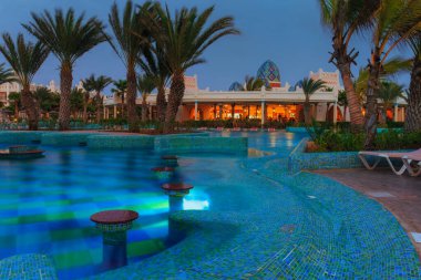Otel havuzundaki zarif mavi ışık, palmiye ağaçlarının silueti ve canlı bir restoran sahnesiyle tamamlanmış sakin bir atmosfer yaratıyor. Gece Vaha 'sı. Yüksek kalite fotoğraf