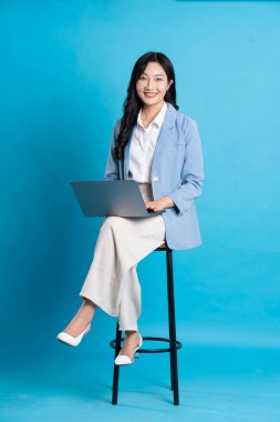 Asyalı iş kadını portresi sandalyede oturuyor, mavi arka planda izole edilmiş.