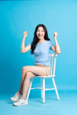 Güzel bir Asyalı kadının portresi bir sandalyede oturuyor, mavi bir arka planda.