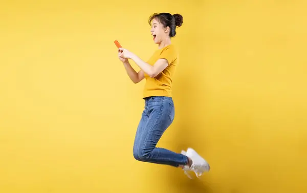 亚洲女孩高飞猛进的全身像照片 手持手机 写着一个新的社交媒体帖子 与蓝色背景隔离 — 图库照片