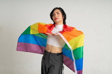 Asyalı eşcinsel bir adamın elinde gökkuşağı bayrağı tutarken beyaz bir arkaplanda güvenle poz vermesi.