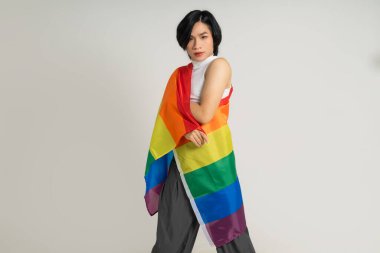 Asyalı eşcinsel bir adamın elinde gökkuşağı bayrağı tutarken beyaz bir arkaplanda güvenle poz vermesi.