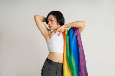 Asyalı eşcinsel bir adamın elinde gökkuşağı bayrağı tutarken beyaz bir arkaplanda güvenle poz vermesi.