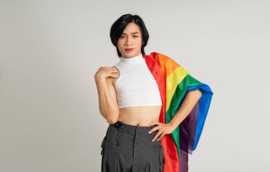 Asyalı eşcinsel bir adamın elinde gökkuşağı bayrağı tutarken beyaz bir arkaplanda güvenle poz vermesi.
