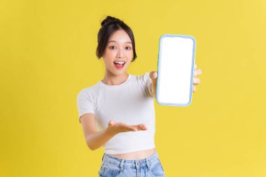 Sarı arka planda neşeli yüzlü genç Asyalı kadın telefonu tutuyor.