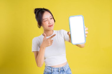 Sarı arka planda neşeli yüzlü genç Asyalı kadın telefonu tutuyor.