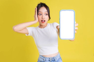 Sarı arka planda neşeli yüzlü genç Asyalı kadın telefonu tutuyor.