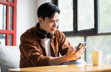 Asyalı bir adamın portresi kafede oturuyor, telefon kullanıyor ve müzik dinliyor.
