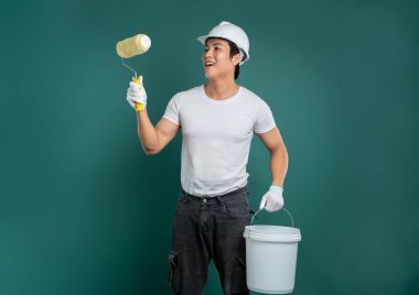 Güvenlik teçhizatı içinde, elinde boya silindiri ve kovayla yeşil bir zemine karşı duran bir ressam. Resim yapmaya hazırlanırken gülümsüyor..