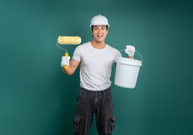 Sert bir şapka, eldiven takmış, elinde boya rulosu ve kovayla boyamaya hazır hevesli bir işçi. Koyu turkuaz arka plan temiz bir stüdyo görüntüsü sağlıyor..