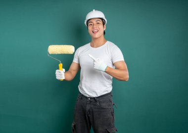 Beyaz bir şapka ve çalışma eldiveni takmış hevesli bir ressam elinde bir boya silindiri tutuyor ve koyu yeşil bir duvara doğru işaret ediyor..