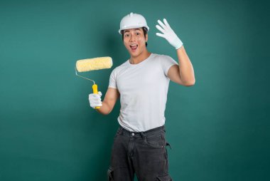 Güvenlik kaskı takan tutkulu bir ressam stüdyoda yeşil arka planda boya silindiriyle el sallıyor. Yenileme veya dekorasyon için hazır, DIY projesi.