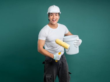 Güvenlik kaskı ve iş giysisi içinde gülümseyen genç bir adam bir boya silindiri ve kova tutuyor, sağlam yeşil bir arka plana karşı, boyamaya ve dekorasyona hazır..