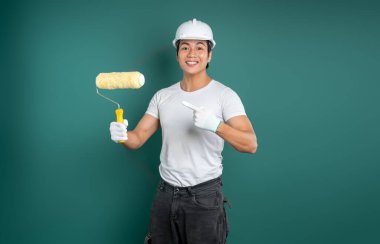 Yeşil bir arka planda boya malzemesi ile gülümseyen bir işçi, bir rulo tutarken eldiven ile işaret ederek, herhangi bir duvara yeni bir görünüm vermeye hazır.