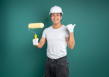 Beyaz tişörtlü ve kot pantolonlu genç adam mavi-yeşil bir duvarın önünde duruyor. Sarı bir silindir tutuyor ve duvardaki boşluğu işaret ediyor..