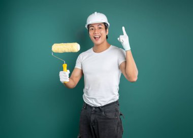 Heyecanlı bir ressam yeşil bir zemine karşı duruyor, kaskı ve eldivenleri var. Bir boya silindiri tutuyor ve yukarı bakıyor, dekore etmeye hazır..