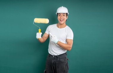 Güvenlik kaskı, beyaz gömlek ve işçi pantolonu giymiş gülümseyen bir ressam yeşil zemine karşı sarı bir boya silindirini işaret ediyor..