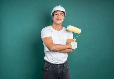 Kasketli, beyaz tişörtlü, kot pantolonlu ve eldivenli bir inşaat işçisi kolları çaprazlanmış bir şekilde duruyor ve deniz mavisi arka planında boya silindiri tutuyor..