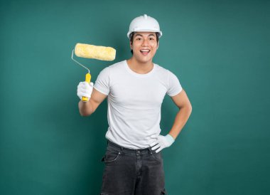 Beyaz şapkalı ve iş eldivenli Asyalı bir adam yeşil arka planda boya silindiri tutuyor. Gülümsüyor, bir resim projesi için hazır..
