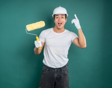 Şapkalı ve eldivenli gülümseyen bir adam boya silindiri tutuyor. Heyecanlı bir ifadeyle yukarıya doğru işaret ediyor, sert yeşil bir zemine dayanıyor.. 