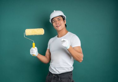 Güvenlik teçhizatı ve koruyucu eldivenleri olan bir ressam elinde boya silindiri tutarken izleyiciye şakacı bir şekilde işaret eder..