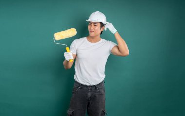 Beyaz tişört, kask ve eldiven giyen bir ressam, deniz mavisi zeminine boya silindiri tutarken yana bakıyor. Resim için hazırlanılıyor.