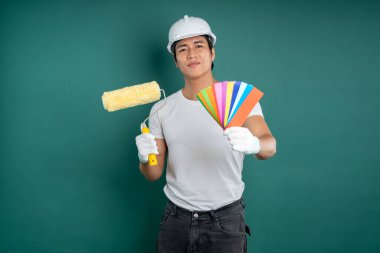 Sert şapkalı Asyalı bir inşaat işçisi yeşil arka planda bir boya silindiri ve renk yelpazesi tutuyor. Profesyonel, DIY ev geliştirme projesi.