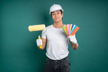 Beyaz gömlekli, kasklı ve eldivenli Asyalı bir adam bir elinde boya silindiri, diğer elinde de yeşil arka planda duran bir palet tutuyor..
