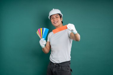 Beyaz bir şapka, eldiven, tişört ve kot giyen gülümseyen bir inşaat işçisi, yeşil arka plan önünde renk örnekleri tutuyor. Stüdyo çekimi.