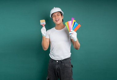 Kasketli ve eldivenli bir ressam gülümsüyor, elinde renk örnekleri ve boya fırçası var. Arka plan tamamen yeşil. Resim servislerine odaklanan bir stüdyo görüntüsü var..