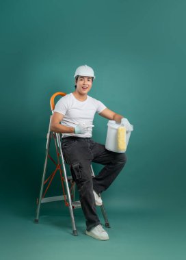 Asyalı tamircinin basamaktaki fotoğrafı. Kasketli ve elinde boya malzemeleri olan. Kovayı işaret ederken gülümsüyor. Yeşil bir arkaplanda izole edilmiş..