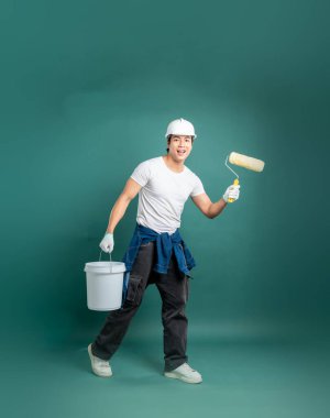 İş kıyafeti giymiş enerjik bir ressam elinde boya silindiri ve kova tutarak, tarafsız yeşil bir zemine gülümsüyor, pozitif ve çalışmaya hazır bir tavır sergiliyor..