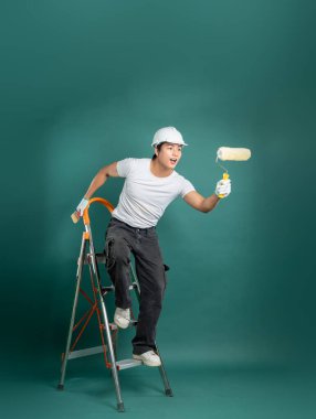 Sert şapka takan bir ressam, elinde boya silindiri olan bir merdivenin üzerinde duruyor. Arkaplan, resmin hareketlerini vurgulayan katı bir renktir..