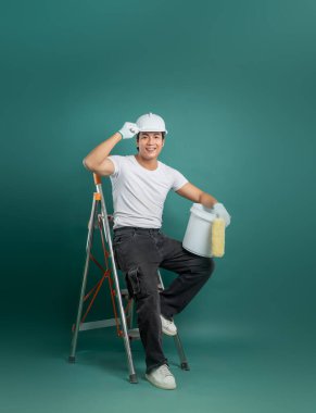 İş kıyafetleri içinde neşeli genç bir adam, beyaz bir miğfer takıyor, elinde boya kovası olan bir merdivende oturuyor, gülümsüyor, turkuaz bir arka planda yenilenmeye hazır..