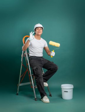 Güvenlik teçhizatı içinde gülümseyen bir ressam elinde boya silindiri olan bir merdivende oturur, yeşil arka planda sağlam bir tabela çizer. Yakındaki kova..